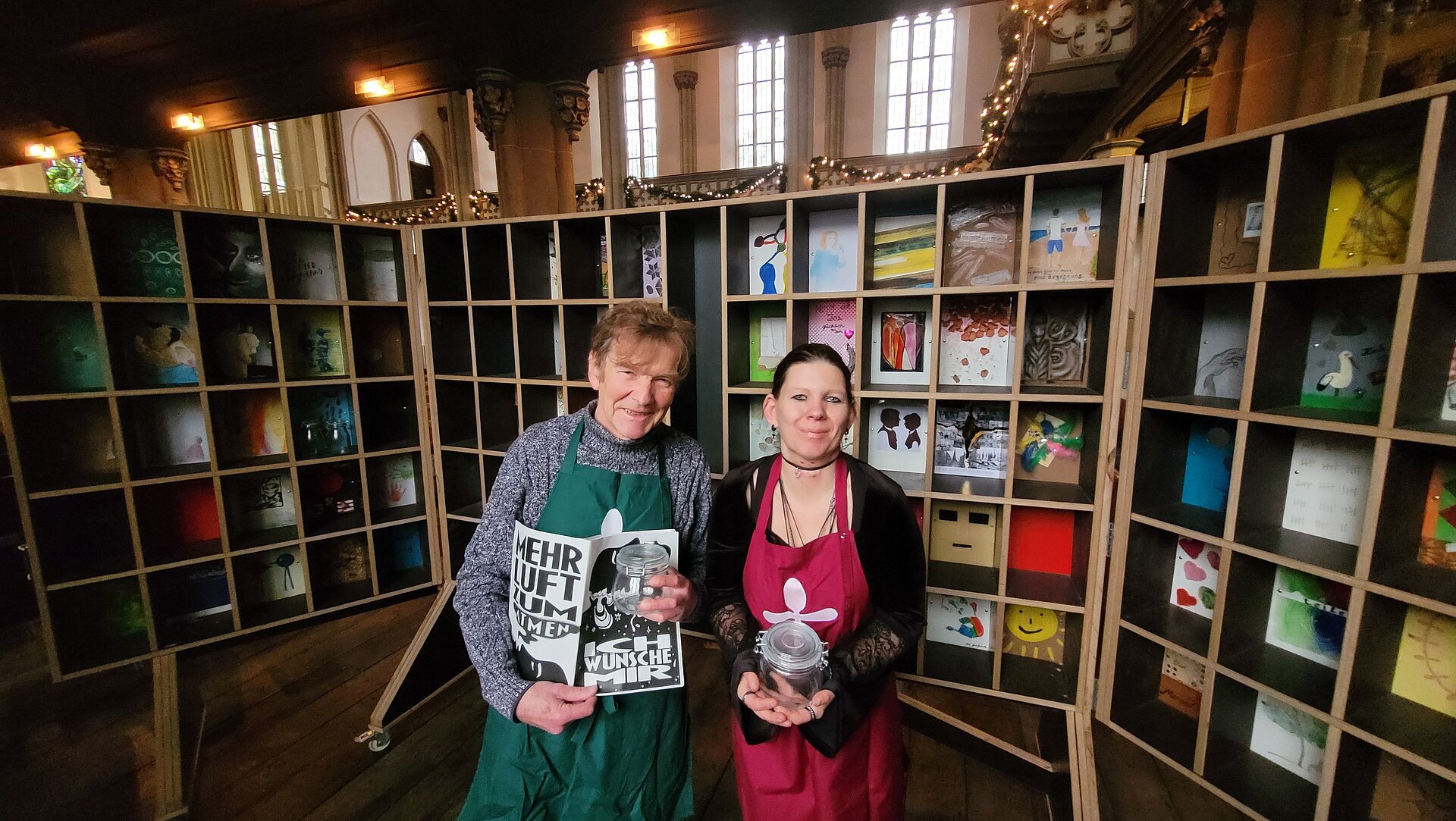 Reinhard Michel und Vanessa Henkelmann begleiten die Kunstaktion. In ihren Händen halten sie Weckgläser, die ab Samstag in der Martin-Luther-Kirche abzuholen sind. Michel und Henkelmann stehen vor dem Regal, das als Suppenregal entstand und im vergangenen Jahr zu einer „Wunschkammer“ wurde.  Aus den Wünschen, die die Gütersloher während der Vesperkirche und im Mai während der „langenachtderkunst“ in dieser Kammer hinterließen, ist ein Heft entstanden, gestaltet von Lara Kees. Gedruckt und gebunden, ist es während der Vesperkirche 2026 zu bekommen.
